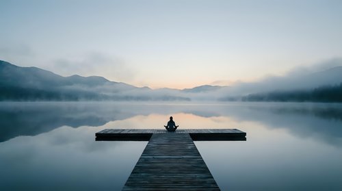 Person sitting alone on wooden dock at misty mountain lake during peaceful sunrise, perfect for meditation and mindfulness content.