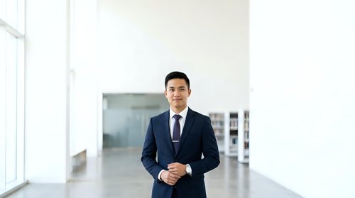 Asian male professional in dark business suit and tie standing in modern office corridor with natural light from windows, suitable for corporate, business, and corporate culture concepts.