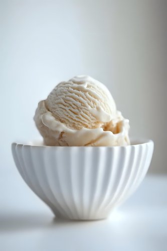 Creamy vanilla ice cream scoop served in elegant white fluted ceramic bowl against light background, minimalist food photography composition for dessert menus.