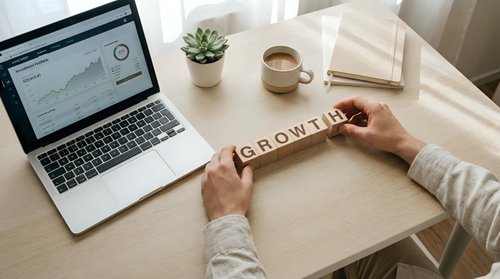 Business professional arranging wooden letter blocks spelling GROWTH on desk with laptop showing analytics, coffee cup and notebook for success planning.