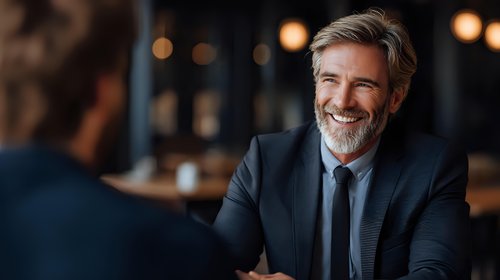 Mature businessman with gray hair and beard wearing dark suit smiling during meeting in restaurant. Professional corporate portrait with warm ambient lighting.