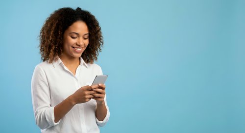 Young African American businesswoman with curly hair smiling while using smartphone on blue background for professional communication and mobile technology.