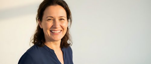 Professional Hispanic female healthcare worker in navy scrubs smiling warmly at camera against neutral background for medical and wellness content.