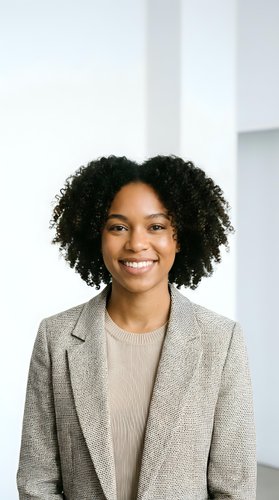 Professional African American businesswoman with curly hair smiling confidently in modern office wearing beige blazer and sweater for corporate headshot.