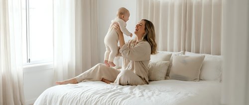Young mother holding up smiling baby in bright white bedroom with natural light streaming through windows creating warm intimate family moment.