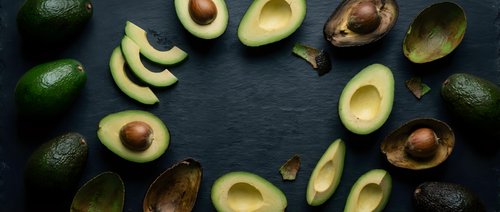 Fresh ripe avocados arranged in circular pattern on dark slate background with halved and sliced pieces showing creamy flesh and pits for healthy cooking.