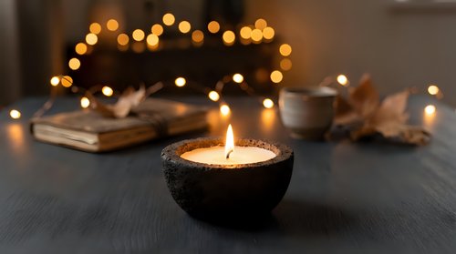 Cozy autumn evening scene with burning candle in dark bowl, warm bokeh lights, old books and dried leaves creating peaceful hygge atmosphere for relaxation.