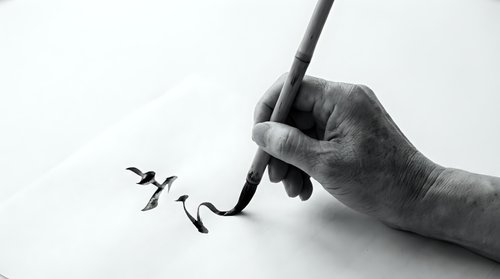 Hand holding brush creating elegant black ink calligraphy strokes on white paper in traditional Asian artistic style for creative design projects.