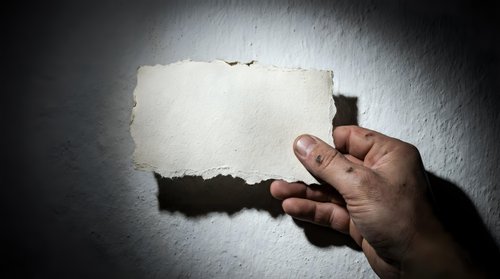 Hand holding blank torn paper sheet against textured wall with dramatic lighting for mockup design projects and creative presentations.