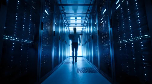 Silhouette of person walking through modern data center corridor with glowing server racks and blue lighting for technology and computing concepts.