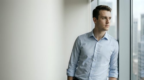 Young Caucasian businessman in light blue shirt standing by office window looking thoughtful and contemplative in modern corporate workspace.