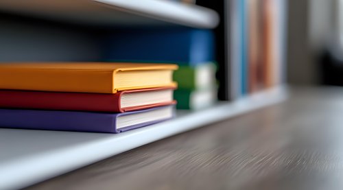 Colorful hardcover books stacked on white shelf against dark wooden table surface with soft blur effect creating minimalist study atmosphere.