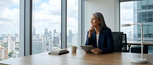 Professional businesswoman in navy blazer sitting at modern office desk with city skyline view through floor-to-ceiling windows during daytime meeting.