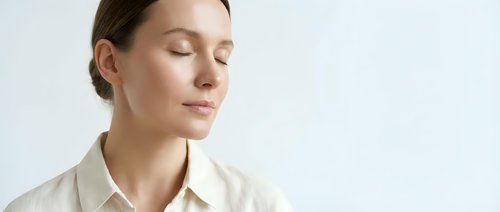 Middle-aged Caucasian woman with closed eyes wearing cream-colored button-up shirt against white background, conveying calm, mindfulness, and peaceful contemplation for wellness and lifestyle content.