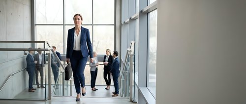 Confident businesswoman walking in modern office corridor with team members in background, corporate leadership and professional workplace environment.