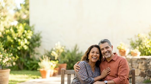 Mature couple embracing on wooden bench in sunny garden setting with blooming flowers and green plants, enjoying outdoor leisure time together.