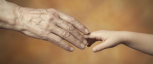 Elderly wrinkled hand gently touching young smooth hand in warm golden light representing intergenerational connection, care, support and family bonds.