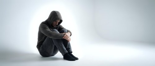 Young man in hoodie sitting alone in contemplative pose against white background expressing sadness, depression, mental health struggles and emotional isolation.