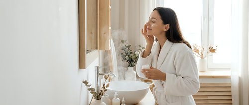 Young Asian woman in white bathrobe enjoying morning skincare routine in bright modern bathroom with natural light streaming through window.