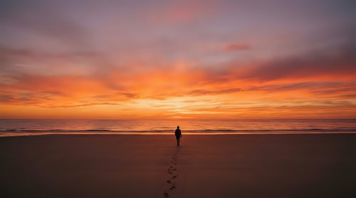 Solitary figure walking on empty beach at sunset with dramatic orange and pink sky reflecting on wet sand creating peaceful meditation moment.