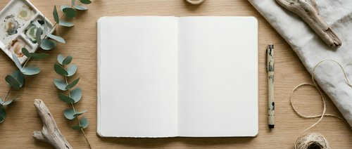 Open blank notebook with pen on wooden desk surrounded by eucalyptus branches, twine, and natural craft supplies for creative writing and planning.