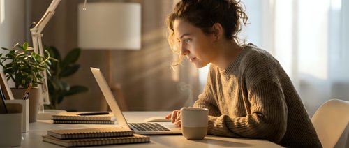 Young woman with curly hair working on laptop at home office desk with coffee cup and plant in warm natural lighting for remote work productivity.