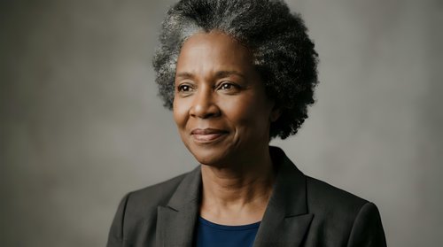 Professional African American businesswoman with natural gray afro hairstyle wearing dark blazer smiling confidently in corporate portrait for leadership concepts.