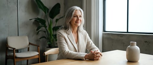 Mature woman with long white hair and fair complexion, wearing a beige blazer, seated at a wooden table in a modern office or home workspace with potted plants and natural light from large windows.