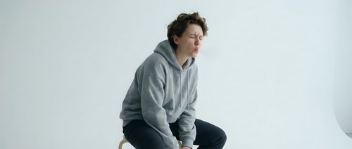 Young Caucasian man in gray hoodie sitting thoughtfully on white surface against minimalist background, expressing contemplation and introspection.