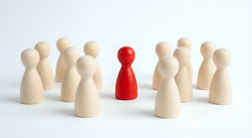 Red wooden pawn figure standing out among group of natural wood pawns on white background. Leadership and uniqueness concept for business presentations.
