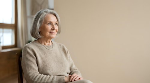 Senior woman with gray hair wearing beige sweater sitting indoors by window, looking away thoughtfully. Portrait for aging, lifestyle, wellness, retirement content.