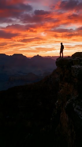 Silhouetted male hiker standing on rocky cliff edge overlooking vast canyon landscape during dramatic golden and orange sunset. Inspiring adventure and exploration concept.