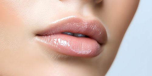Close up of plump, glossy natural pink lips on light skin tone female face showing professional makeup and skincare results against soft focus background.
