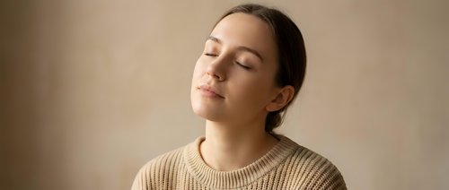 Young woman with closed eyes in peaceful meditation pose wearing beige knit sweater against neutral background for wellness and mindfulness concepts.