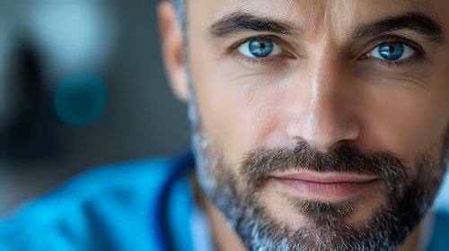 Close up portrait of mature Caucasian man with bright blue eyes, dark beard and gentle smile looking directly at camera against blurred background.