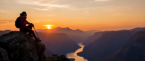 Hiker sitting on mountain peak at sunrise overlooking winding river valley below, contemplating scenic landscape during golden hour adventure and exploration.