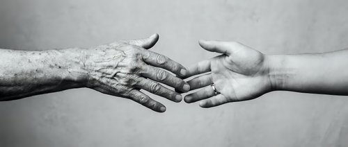 Young and elderly hands reaching toward each other in black and white, symbolizing intergenerational connection, care, and human bond across age groups.