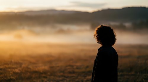 Silhouette of young woman with curly hair standing in misty field during golden hour sunrise. Peaceful morning landscape with rolling hills and warm sunlight.