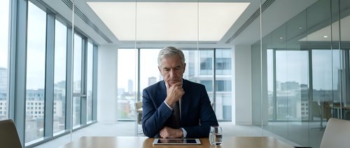 Mature male executive in dark suit sitting at conference table in modern high-rise office with city skyline views, contemplative expression, professional business environment.
