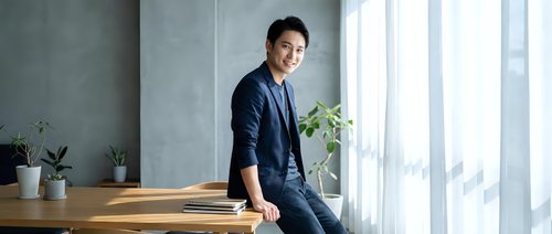 Young Asian businessman in navy suit standing confidently at modern office desk with plants and large windows, professional corporate environment.