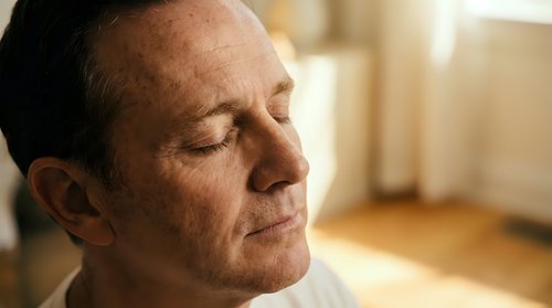 Middle-aged man with light skin tone and brown hair in profile, eyes closed in contemplation or meditation, warm indoor lighting, suitable for wellness and mindfulness concepts.