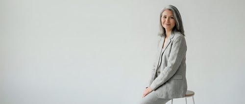 Professional mature woman with gray hair in light blazer sitting confidently on white stool against neutral background for business presentations.
