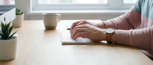 Professional woman typing on laptop computer at modern wooden desk workspace with coffee mug and plants creating productive home office environment.