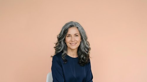 Middle-aged woman with long wavy gray hair and warm smile wearing dark navy blue top against soft peachy beige background. Portrait suitable for business, healthcare, corporate websites, and professional profiles.