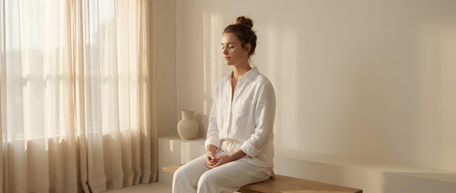 Young Asian woman in white pajamas sitting peacefully on bed in bright bedroom with natural sunlight streaming through curtains creating serene morning atmosphere.
