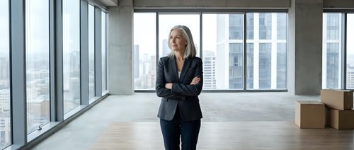 Middle-aged Caucasian female businesswoman in dark blazer standing in modern empty office space with floor-to-ceiling windows overlooking city skyline. Suitable for corporate, real estate, and business concepts.
