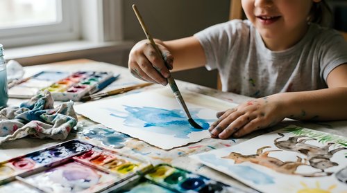 Young Asian girl painting watercolor artwork at home table with brushes and art supplies for creative education and artistic development.
