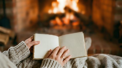 Unrecognizable person in cozy sweater holding open book near fireplace, enjoying reading by warm fire during winter relaxation and leisure time.