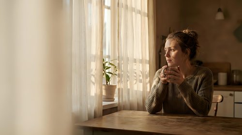 Young Caucasian woman in cozy sweater sitting at wooden table by window with warm sunlight, holding coffee mug in peaceful morning kitchen interior.