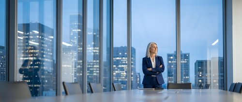 Confident blonde businesswoman standing in modern corporate boardroom with arms crossed overlooking illuminated city skyline through floor-to-ceiling windows at dusk.
